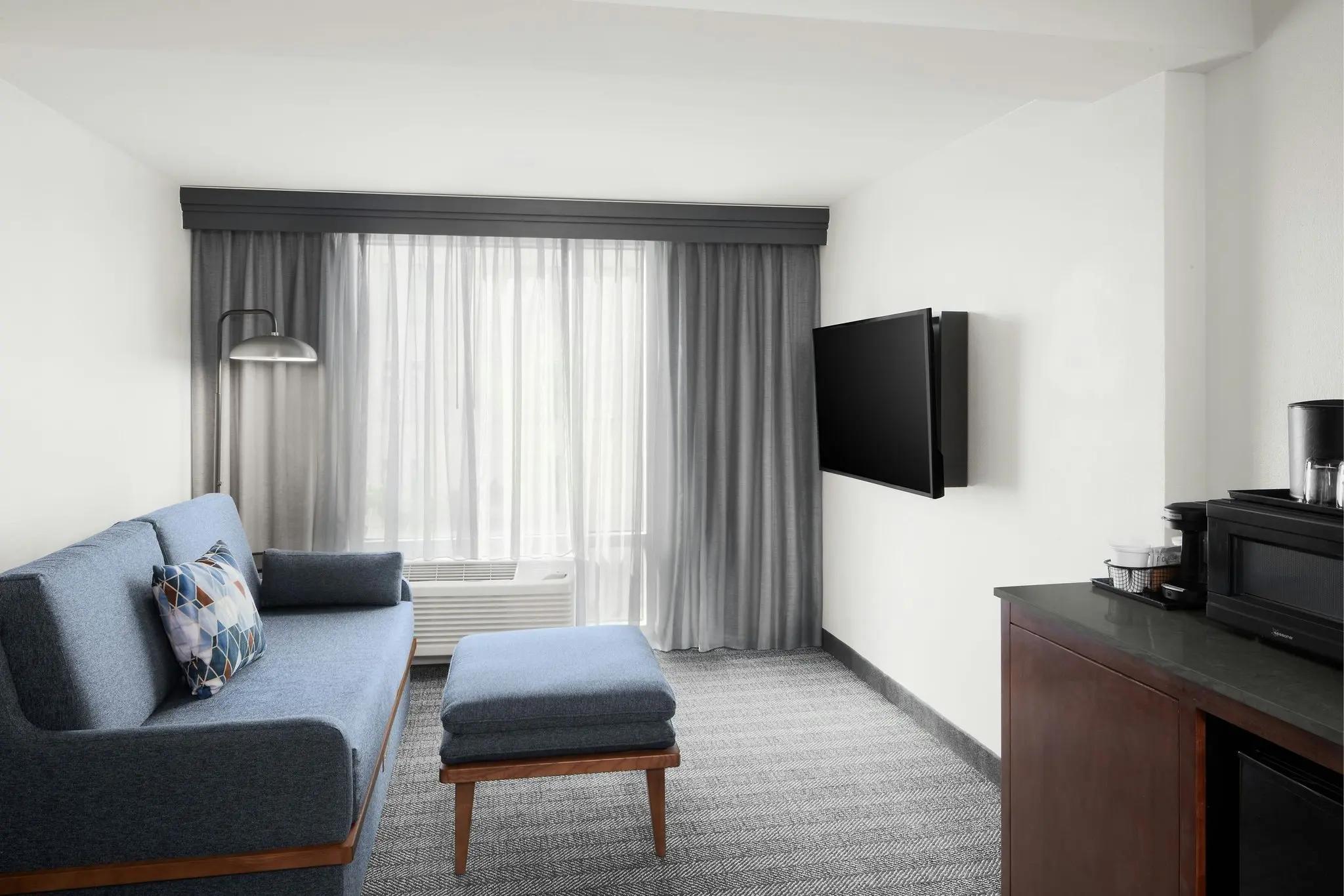 A modern hotel room seating area featuring a blue sofa, ottoman, flat-screen TV, and coffee station.