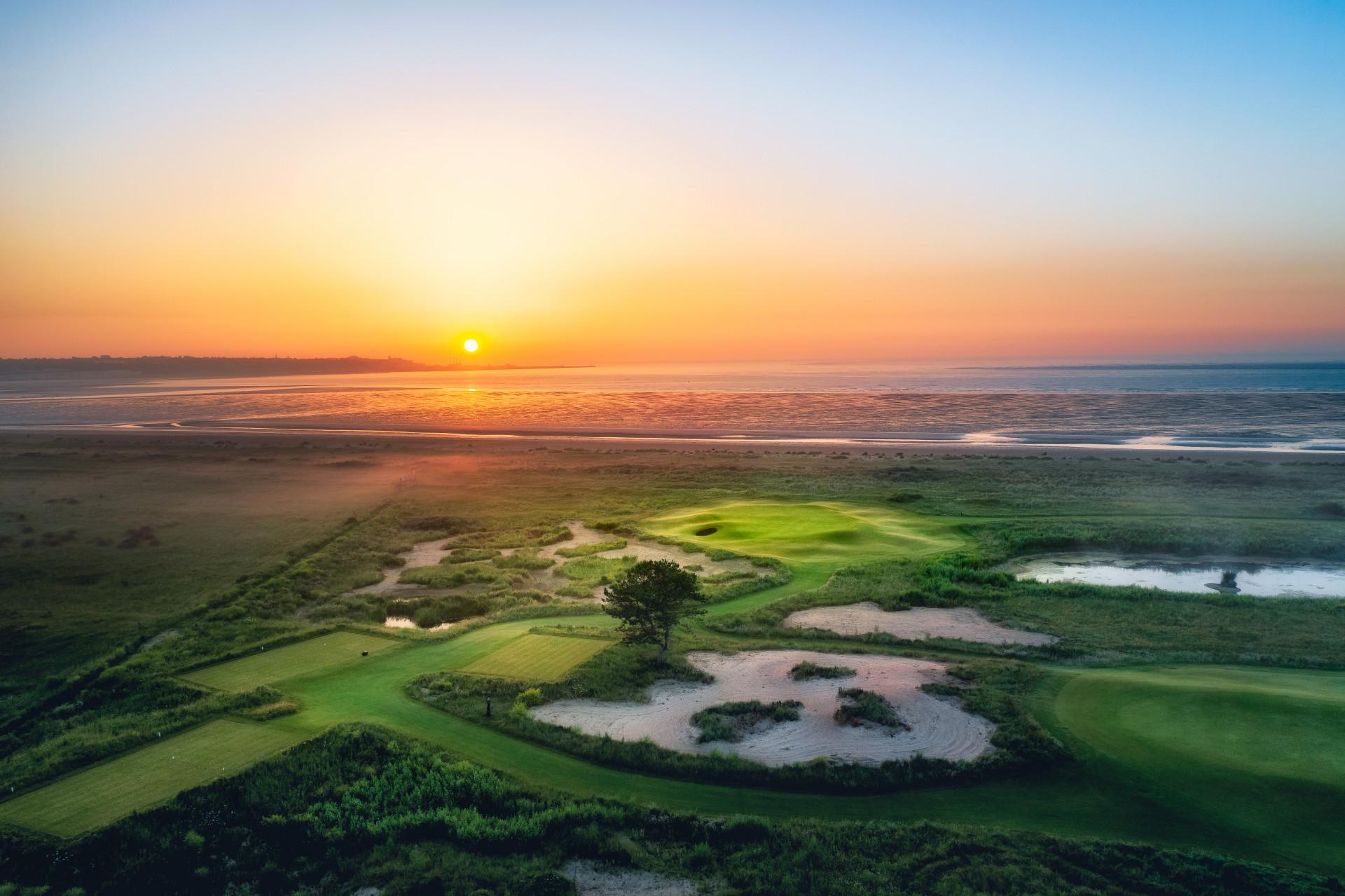 Sunrise over a coastal green with sand bunkers and calm waters.