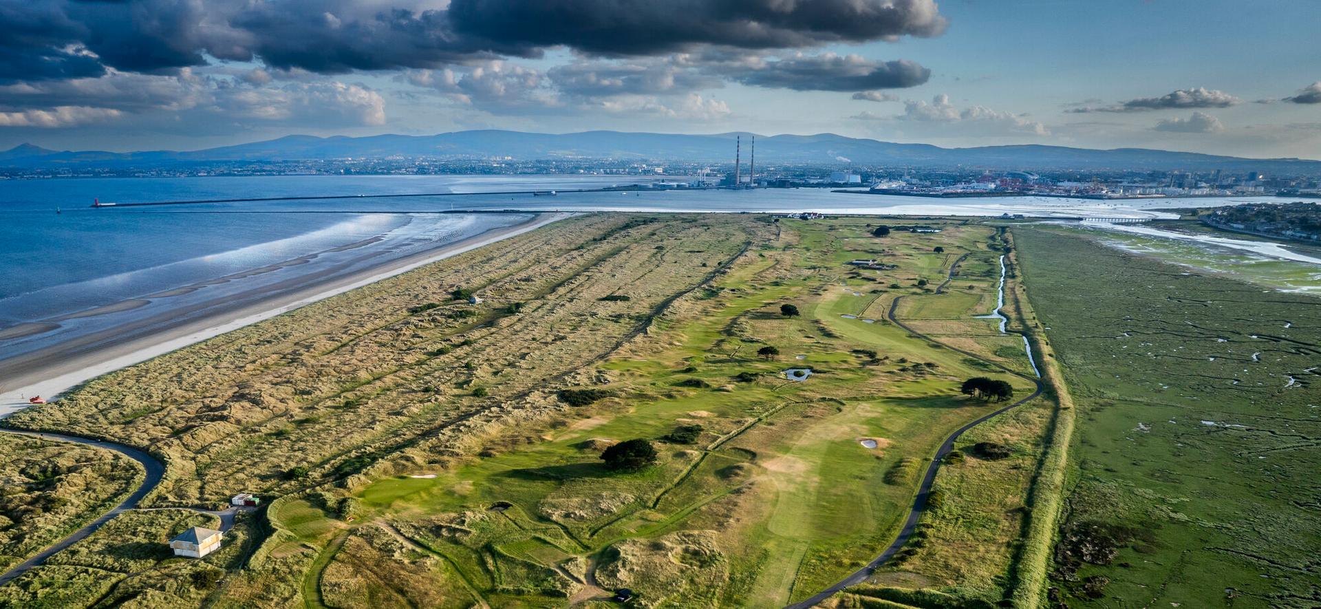 An aerial view of a scenic golf course stretching along the coastline with mountains and a city skyline in the background.