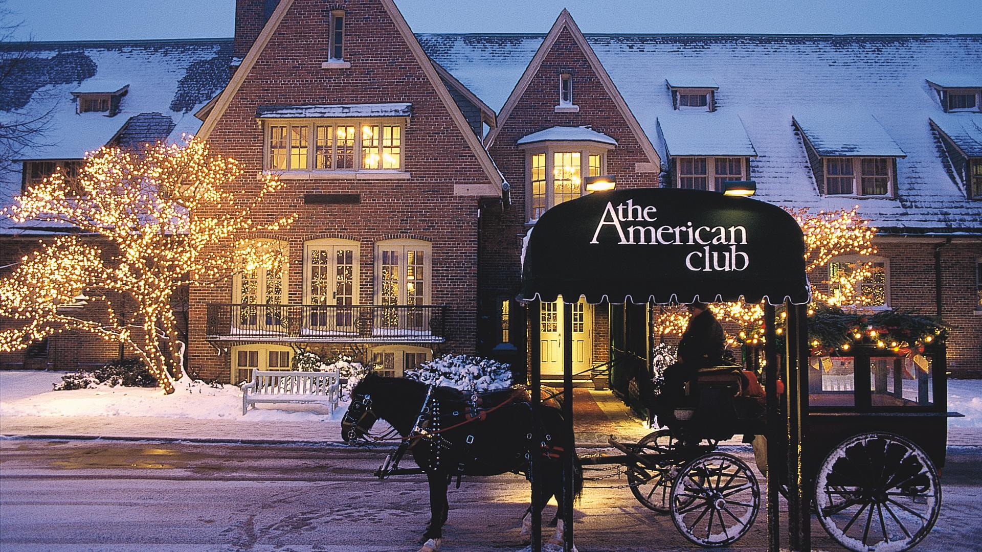 A charming, snow-covered exterior of The American Club with festive lights and a horse-drawn carriage.