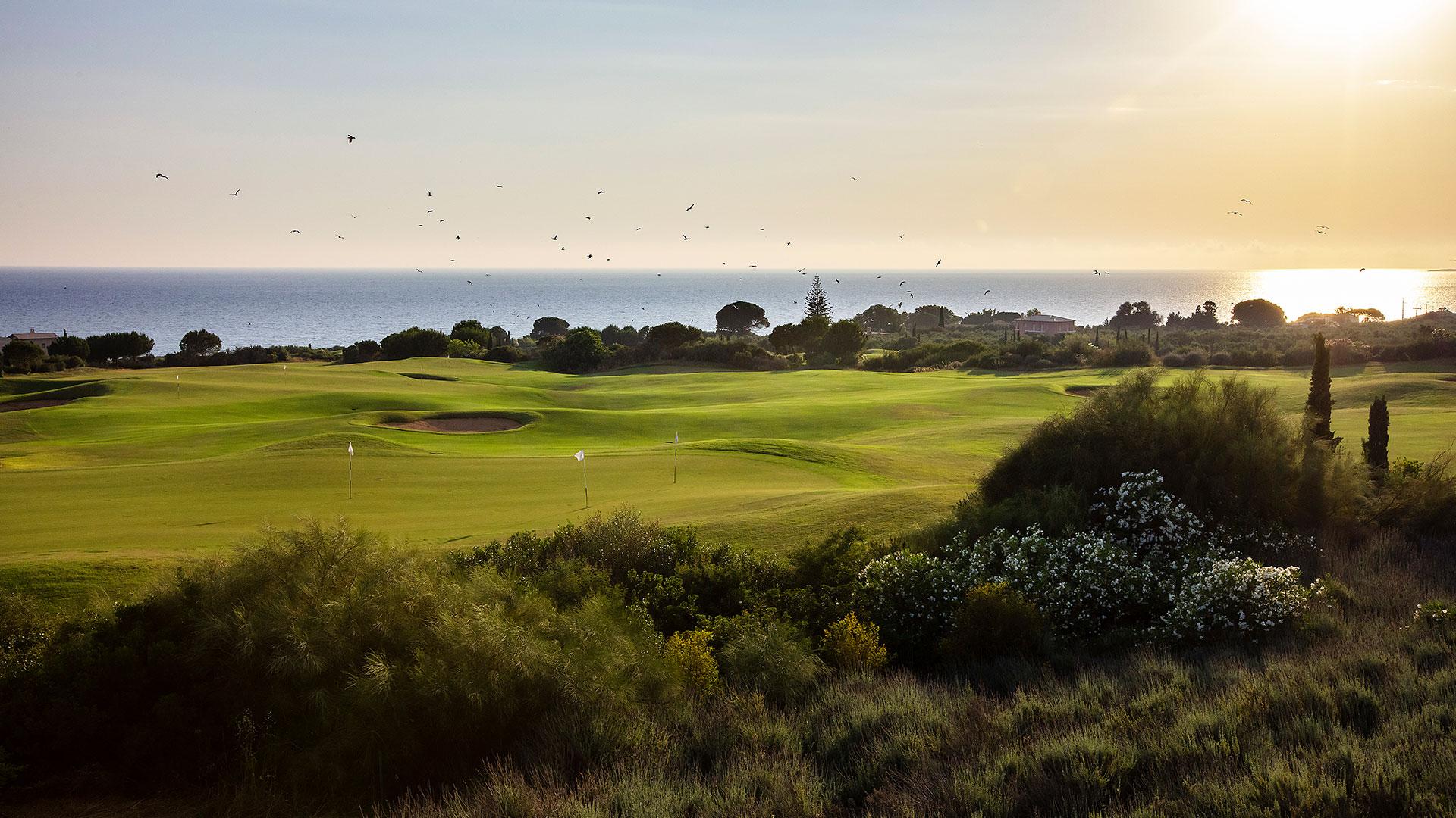 A lush golf course near the sea is surrounded by trees and houses under a golden sky.