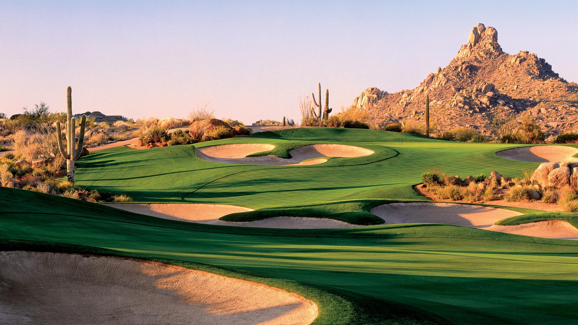 A lush golf course hole framed by desert cacti and rugged mountains at sunrise.