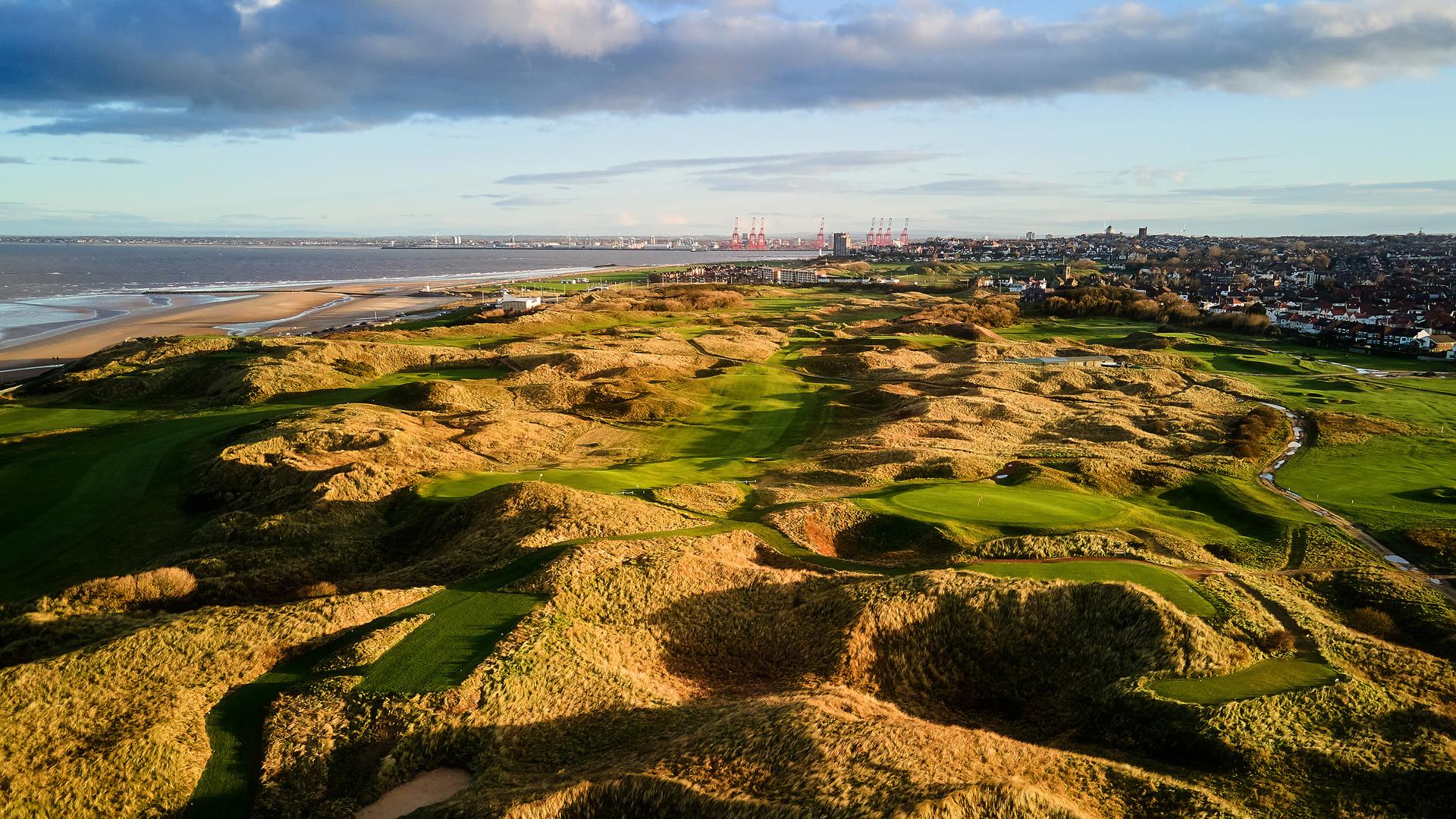 A panoramic view of a golf course with rugged dunes and a nearby town on the horizon.