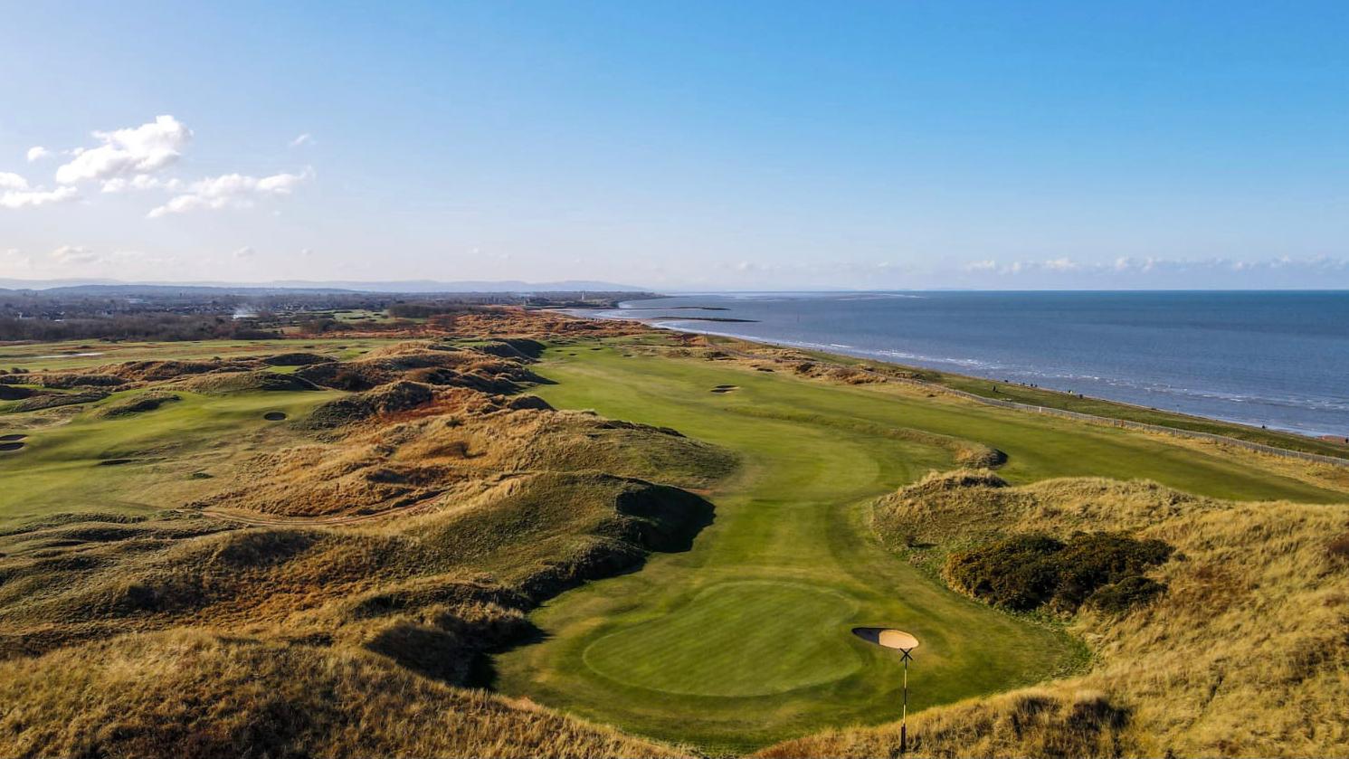 A stunning golf course stretches along the coastline with rolling dunes and a clear blue sky.