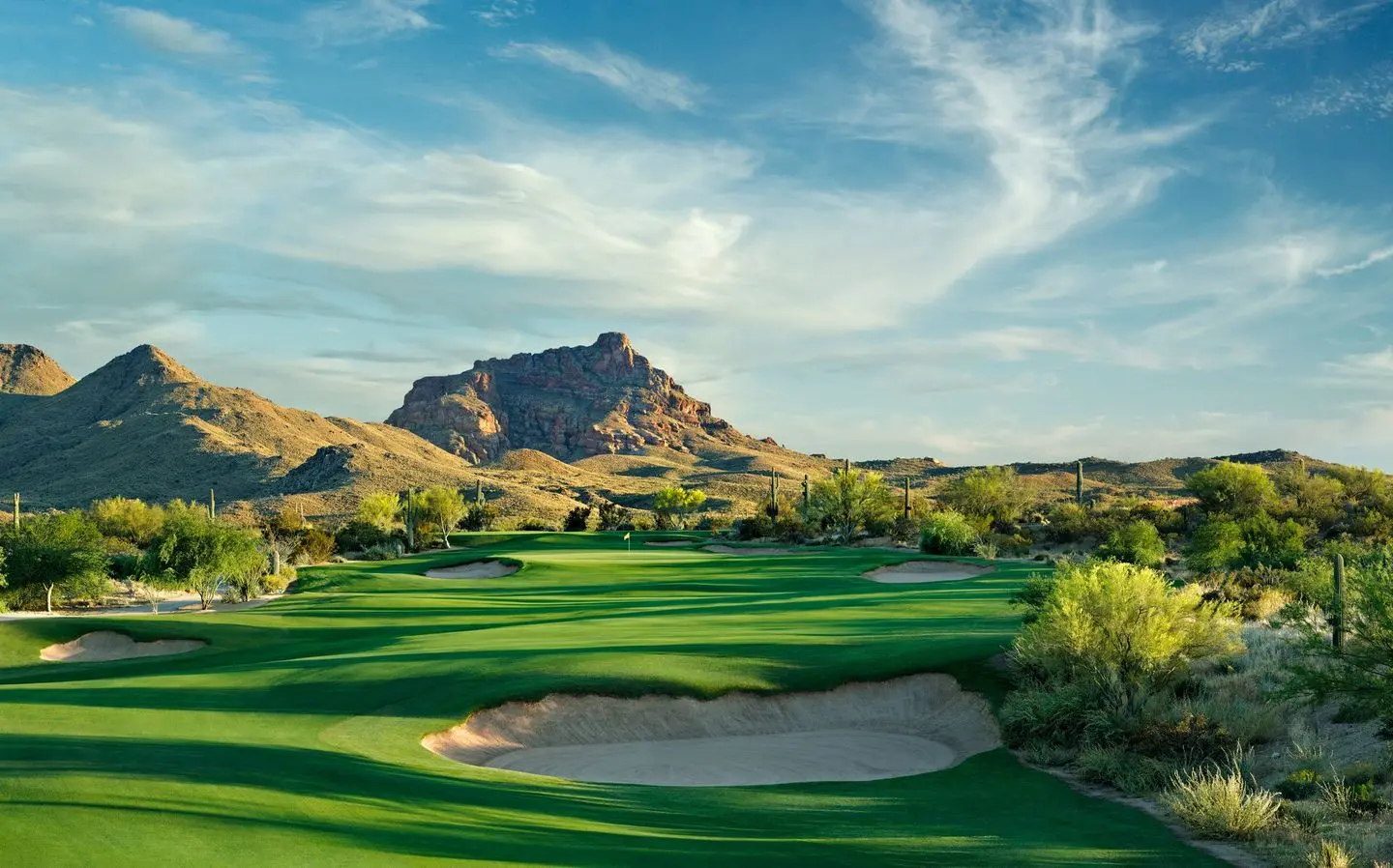 A lush desert golf course with sand bunkers and rugged mountains in the background.