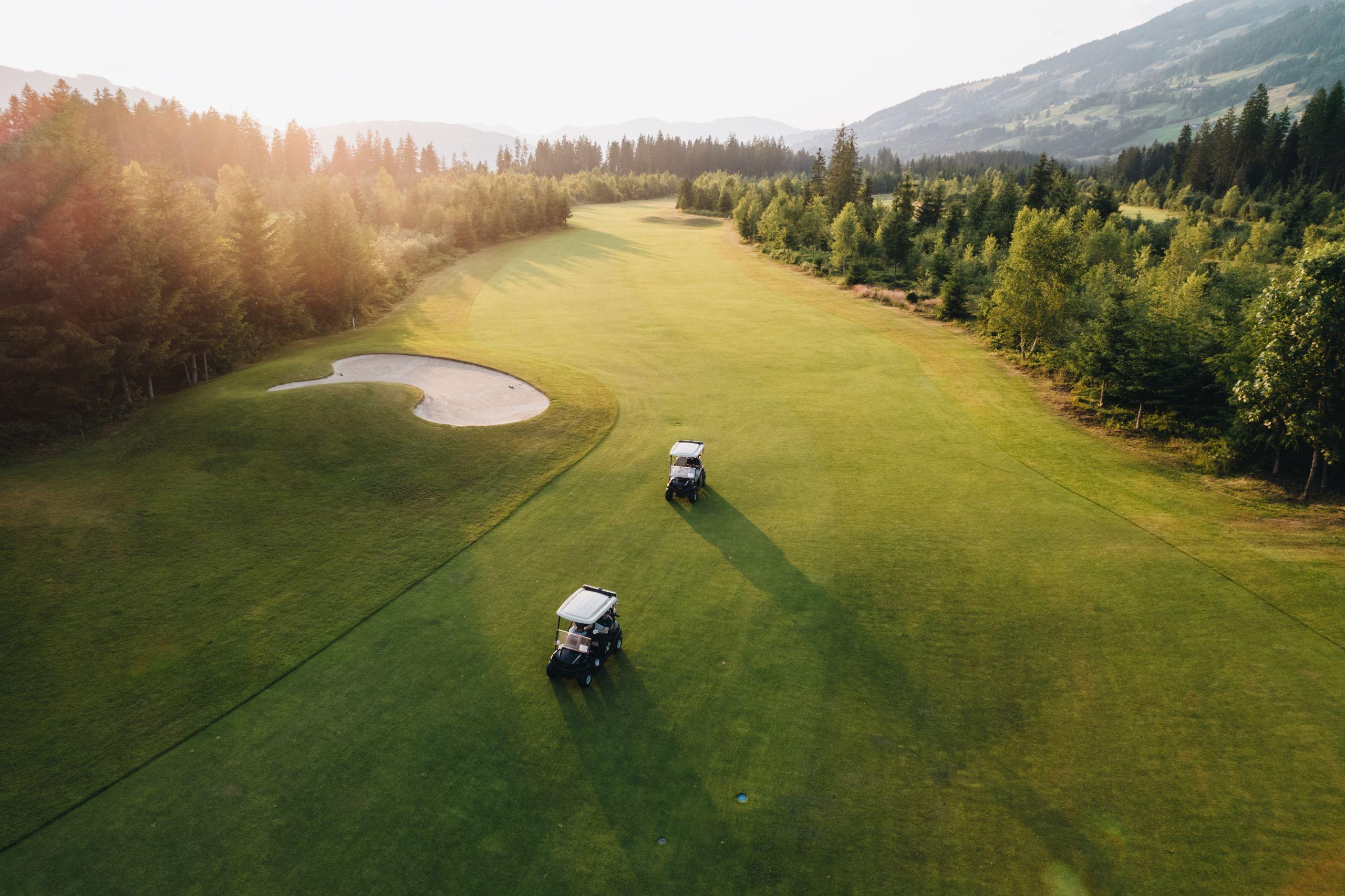 Two golf carts driving along a scenic fairway surrounded by trees and mountains.