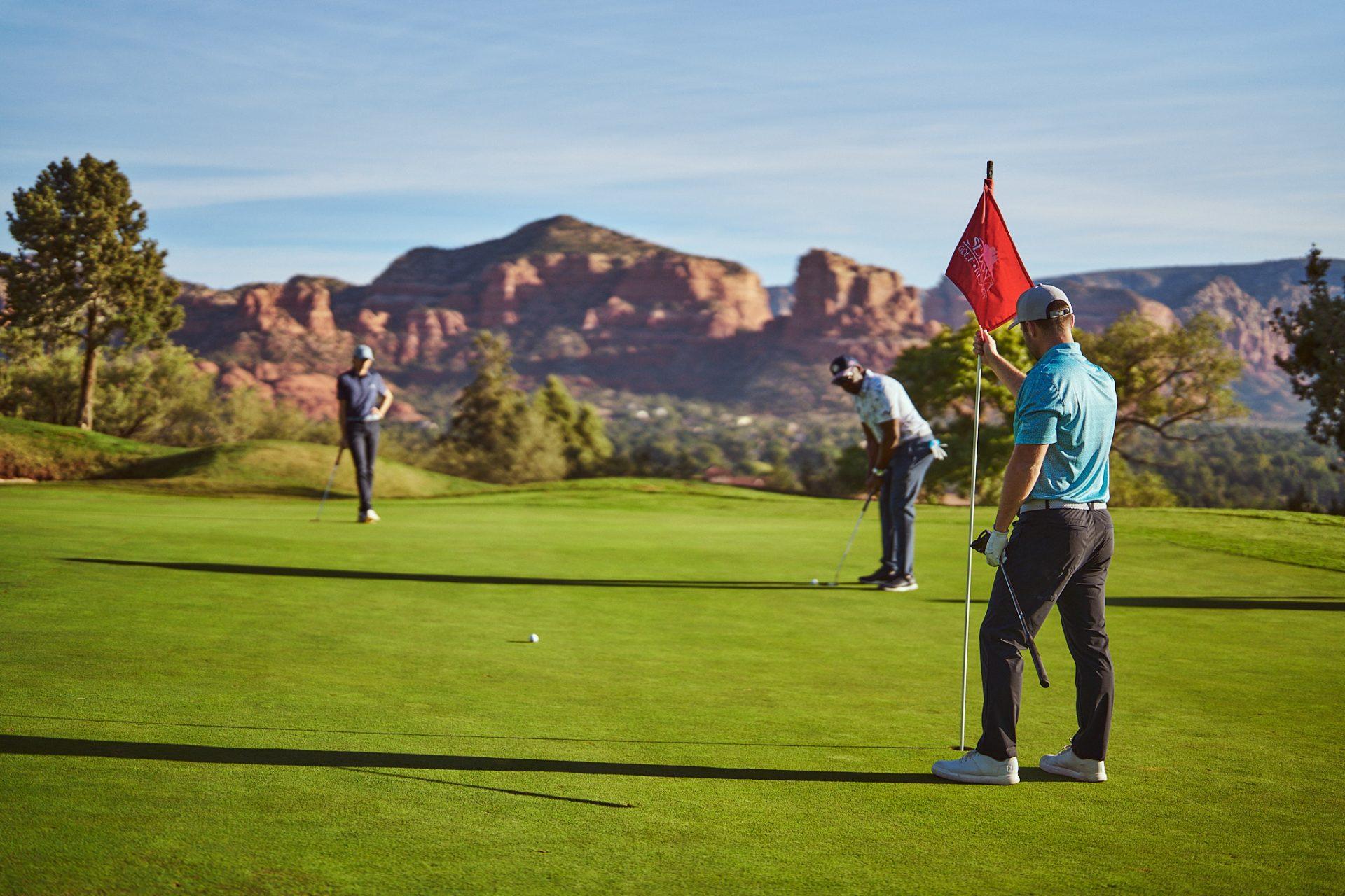 Golfers putting on a manicured green with a red branded flagstick and mountains in the distance
